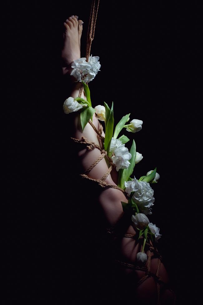 a captivating image showcasing the art of shibari.  a woman's legs and feet are intricately bound with naturalcolored rope, adorned with lush white tulips and greenery woven throughout the bondage. the dark background provides a stark contrast, highlighting the delicate details of both the ropework and the flowers. the overall mood is artistic and sensual, with a focus on the interplay of textures and the elegant flow of the rope and flowers around the body. the image is meticulously composed, suggesting a deliberate artistic approach rather than explicit content.