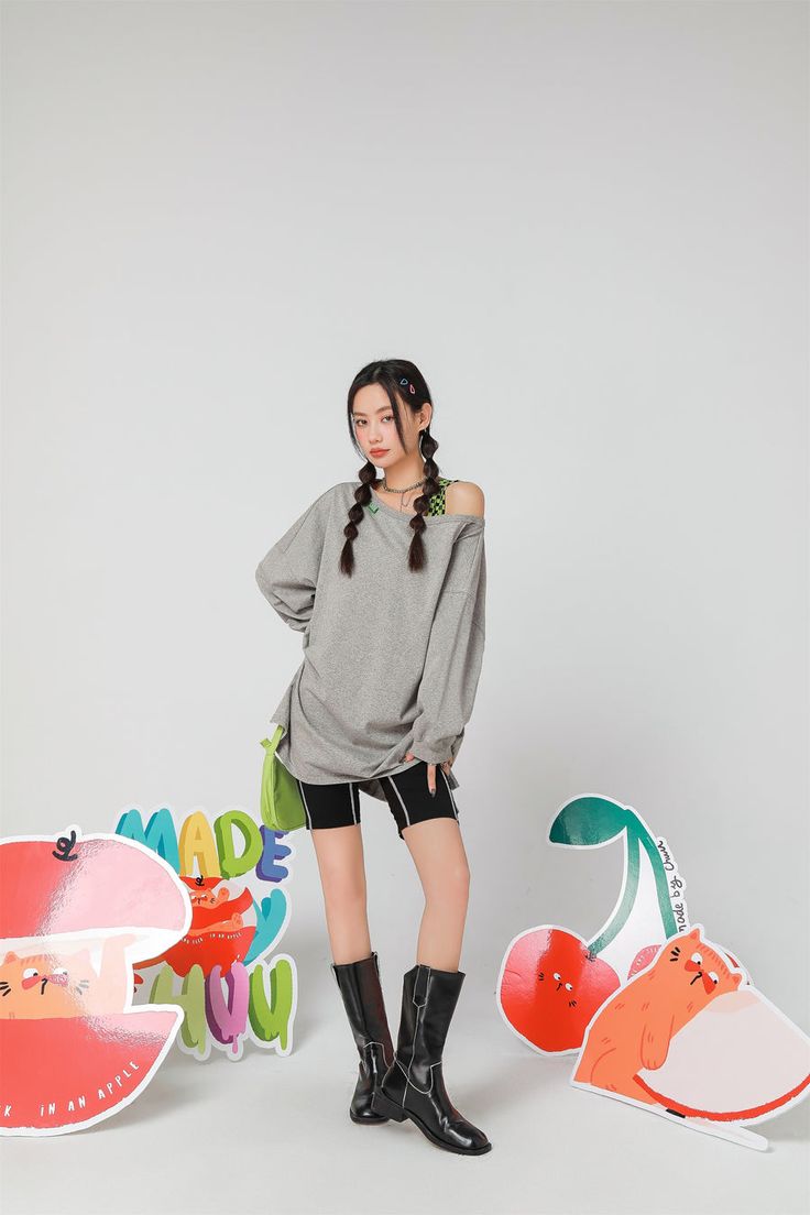 a young woman with long braids is standing in a studio, posing against a backdrop of colorful cartoon cutouts of apples and cherries.  she's wearing an oversized gray sweatshirt, black shorts, and black ankle boots.  her overall style is playful and cute. the backdrop elements suggest a fun and youthful brand or product.