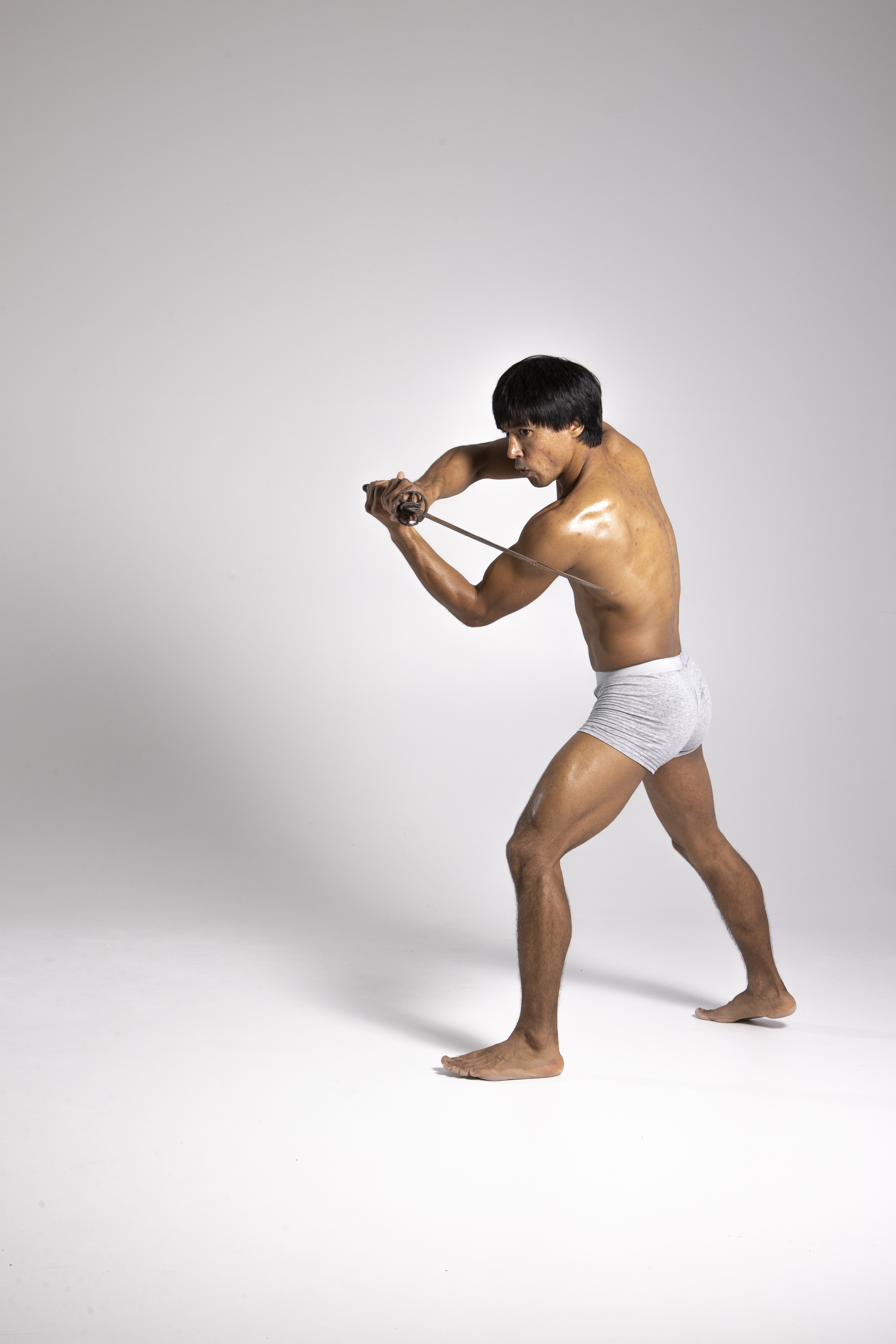 a shirtless, muscular asian man in underwear is wielding a sword in a studio setting. he is positioned in a side view, barefoot, with oiled skin, demonstrating a samurai stance. the man appears to be engaged in martial arts or sword practice.