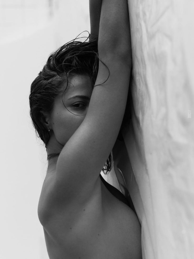 the black and white image shows a woman's upper body, from the back and slightly angled to the side, leaning against a lightcolored wall. her hair is wet and pulled back from her face, which is mostly obscured by her arm. the overall mood is sensual and intimate, with a focus on the textures and curves of the woman's body. the minimalist composition enhances the feeling of mystery and elegance.