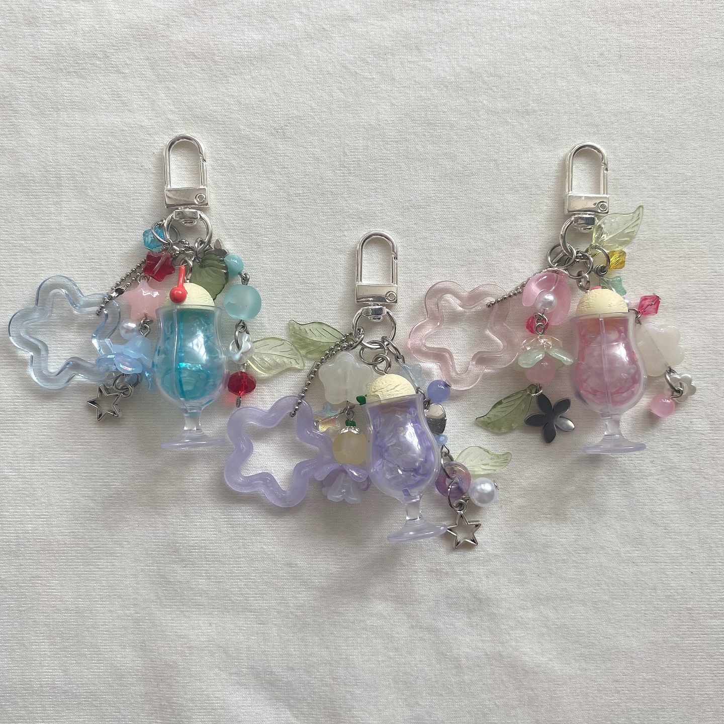 the image shows three keychains laid on a white surface. each keychain features a silvertoned metal clip attached to a collection of charms. the charms include pastelcolored cloud outlines (blue, purple, and pink), cocktail glassshaped charms filled with colored liquid (blue, purple, and pink), and various beads, flowers, leaves, and star charms in complementary colors. the keychains appear to be handmade, showcasing a playful and whimsical design.