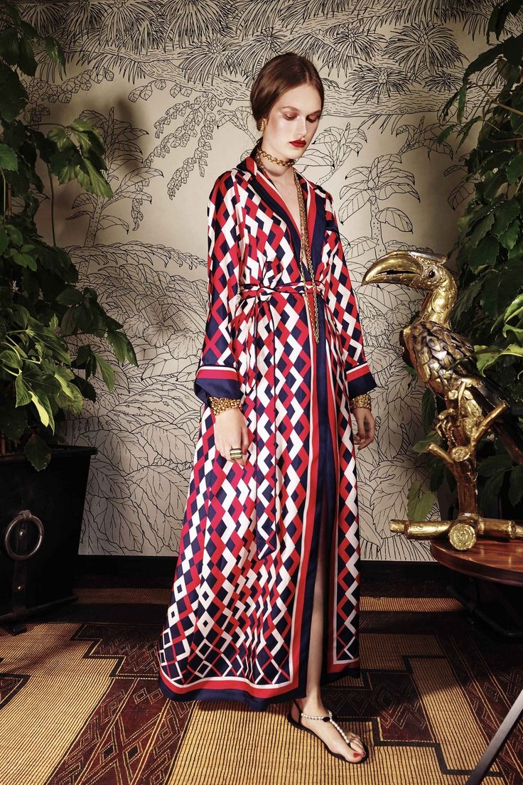 a fulllength shot of a woman wearing a striking red, white, and blue geometricpatterned silk maxi dress. the dress has a kimonostyle silhouette with long sleeves and a belted waist. she is accessorized with gold jewelry, including a choker necklace and bracelets, and simple gold sandals. the model is standing in a room with a rich tropicalthemed wallpaper featuring palm trees and other foliage. a gold toucan statue is visible to her right, and lush potted plants flank either side of her. the floor is covered in a textured rug with warm tones. the overall mood is elegant and sophisticated.