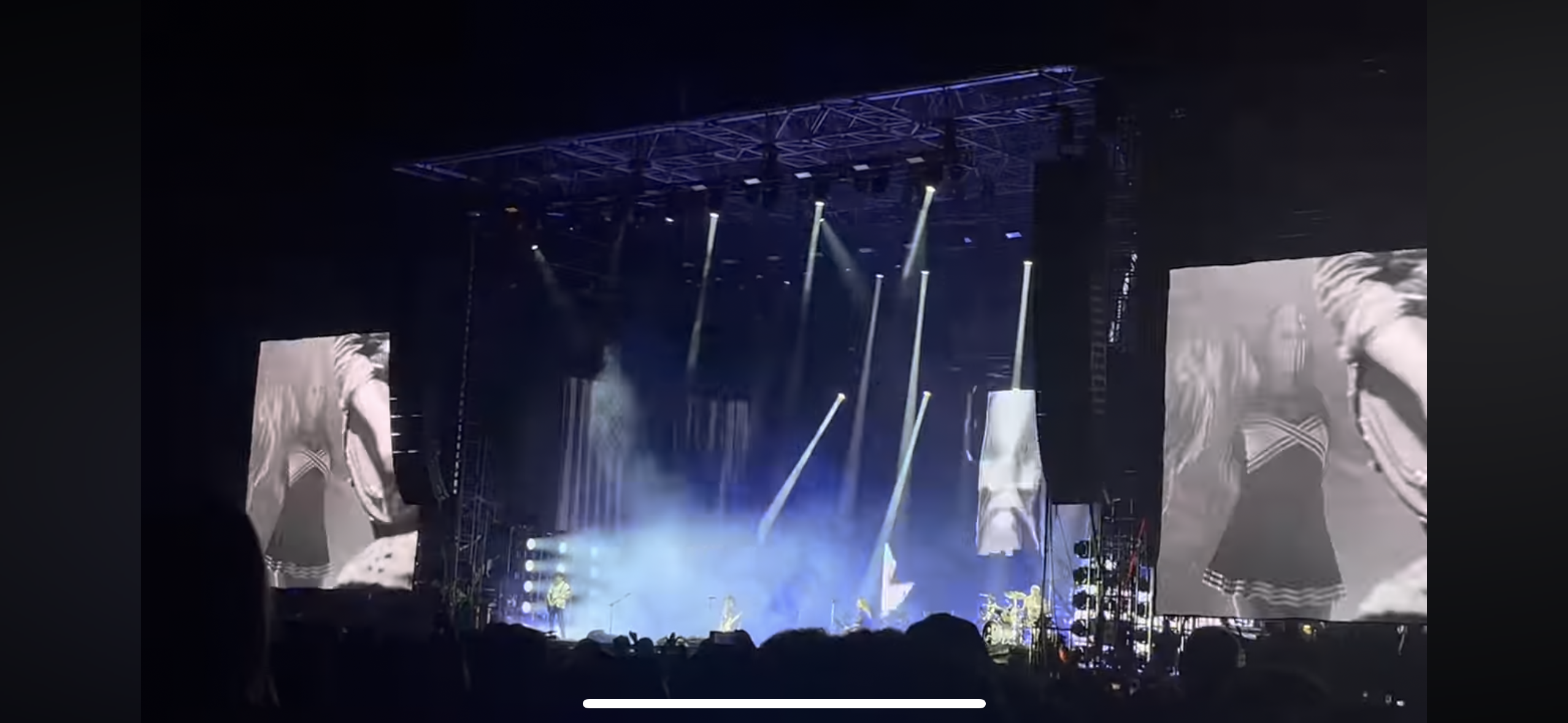 this is a photo of a concert performance.  there is a large stage with a band performing on it.  they are playing instruments and singing into microphones, while the audience watches from below.  the stage is lit up with many spotlights, and smoke machines are being used, creating a dramatic and exciting atmosphere. the two large screens on either side of the stage play videos, and one shows a woman in a skirt with a black and white checkered pattern.