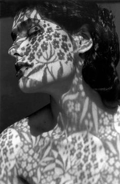a black and white closeup portrait of a woman, her face and upper body adorned with intricate shadow patterns created by sunlight filtering through a lace or patterned fabric.  the shadows create a floral and leafy design across her skin, resulting in a surreal and artistic effect. the woman's expression is serene, and her pose is contemplative. the overall mood is one of beauty, mystery, and artistic expression.