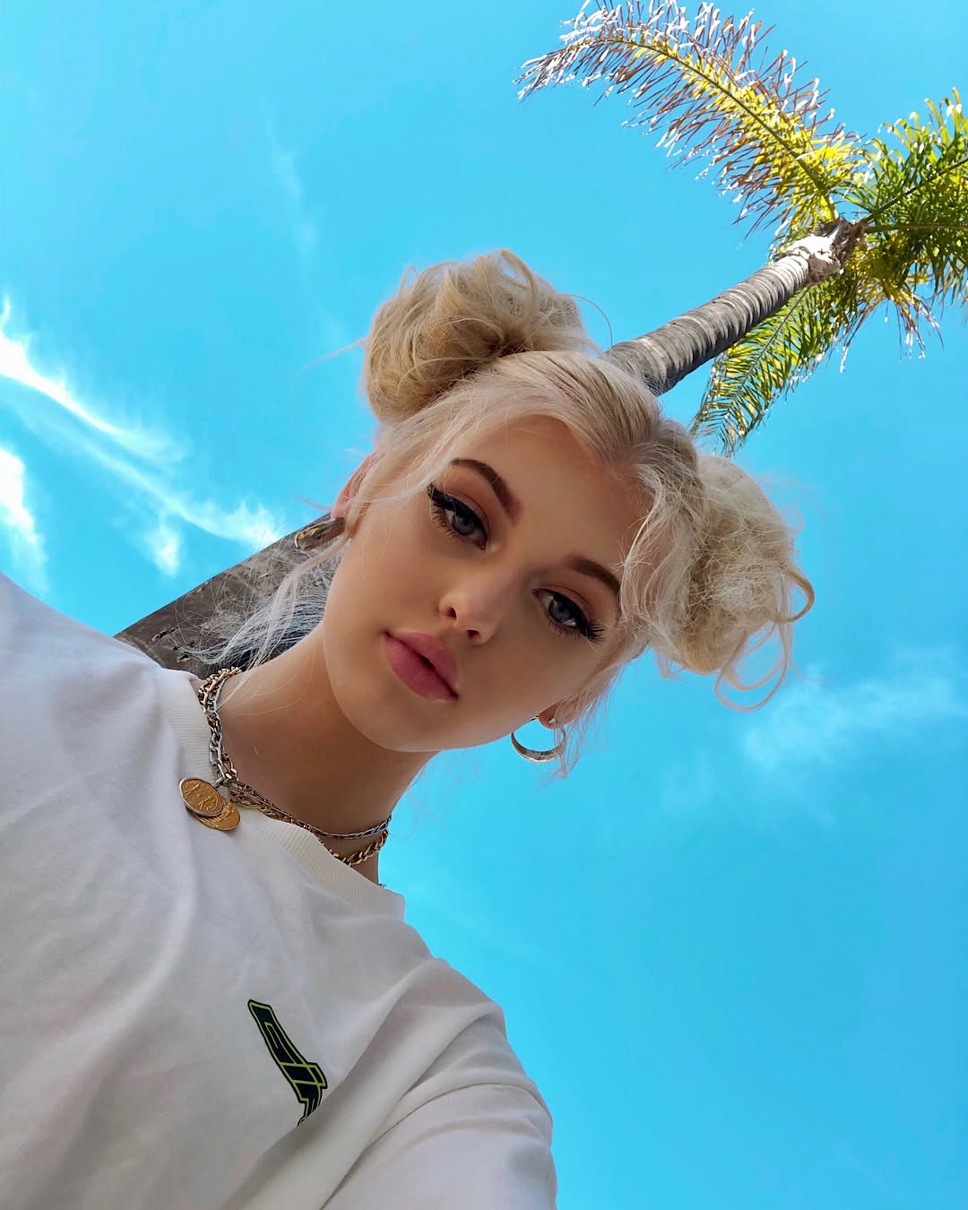 a woman with blonde hair styled in space buns takes a selfie from a low angle, looking up at the camera. she's wearing a white tshirt, necklace, and earrings. behind her is a bright blue sky and a palm tree.