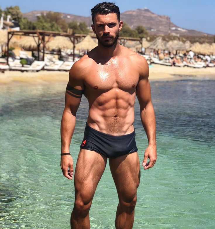 a picture of a muscular, tanned man standing in shallow ocean water.  he is wearing black swim trunks and has a small tattoo on his left arm.  he is facing the camera and looking directly at the viewer. the background is a sunny beach scene in greece.