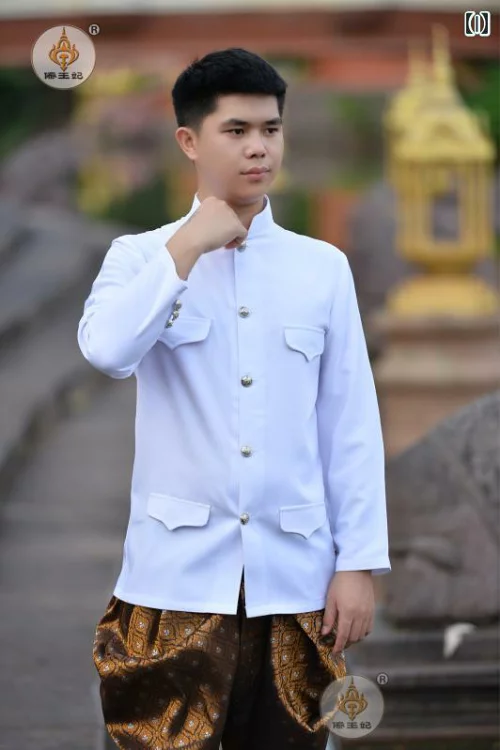 a man in a white traditional thai shirt and gold patterned pants stands smiling in front of a temple.  the temple in the background is slightly out of focus.  the man is dressed in traditional thai attire.