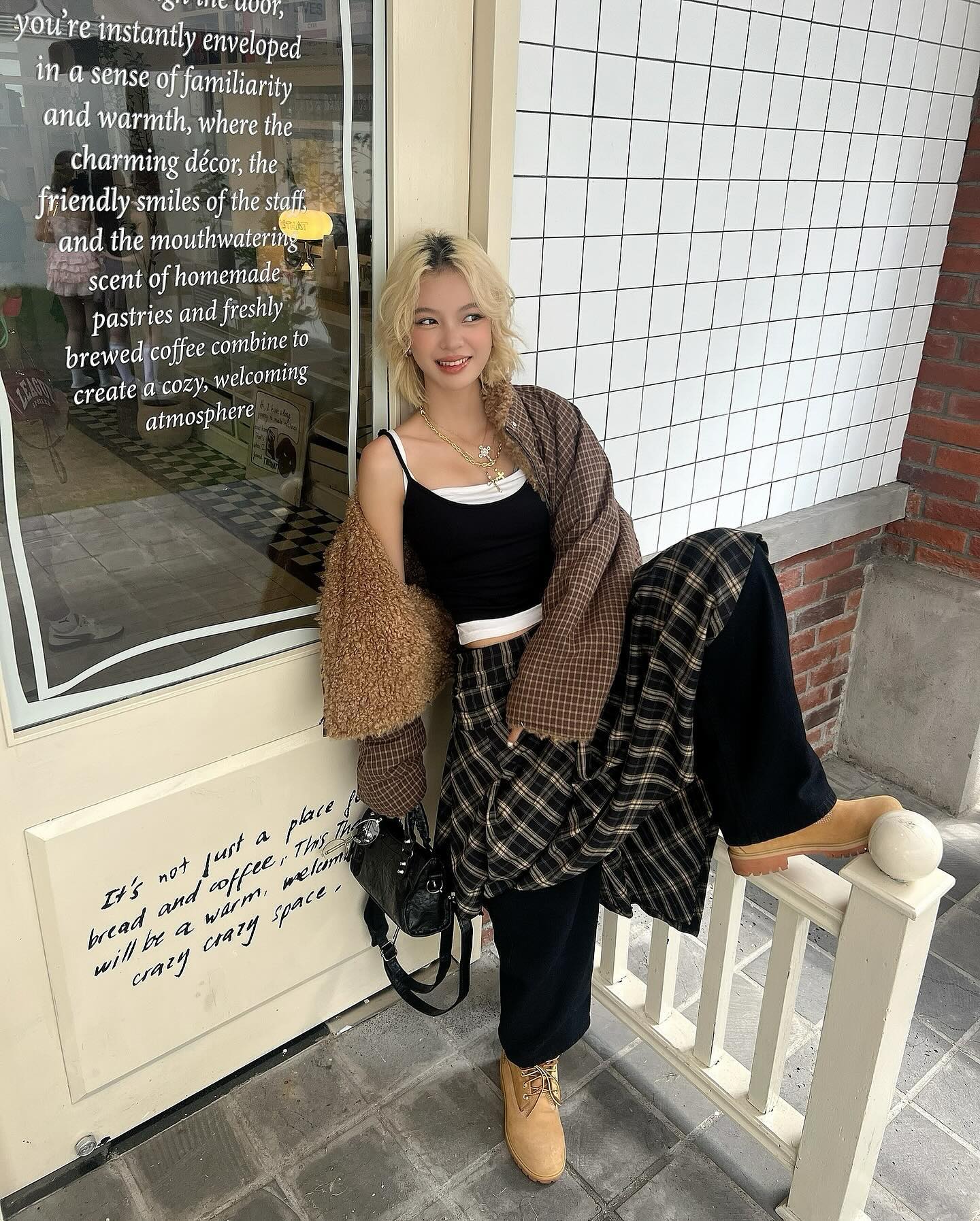 the image shows a young woman standing outside a coffee shop. she is wearing a stylish outfit that includes a plaid skirt, black top, brown jacket, and timberland boots. the coffee shop's sign features text describing a warm and welcoming atmosphere. the overall aesthetic is cozy and trendy, with falllike colors and textures.  the woman is positioned next to a white railing near the coffee shop's entrance.