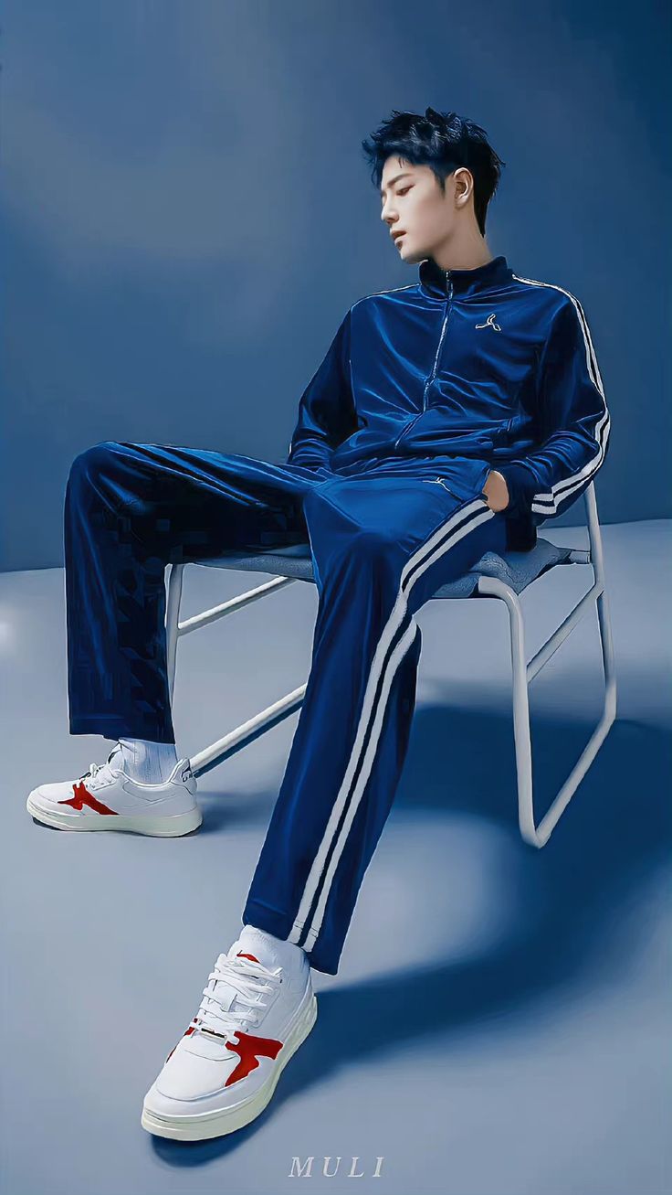 young man in a blue tracksuit and white sneakers sits on a white metal chair against a blue backdrop in a studio setting. he poses with one leg extended, showcasing the outfit and footwear. the lighting is soft and artificial, creating a contemporary and stylish ambiance.