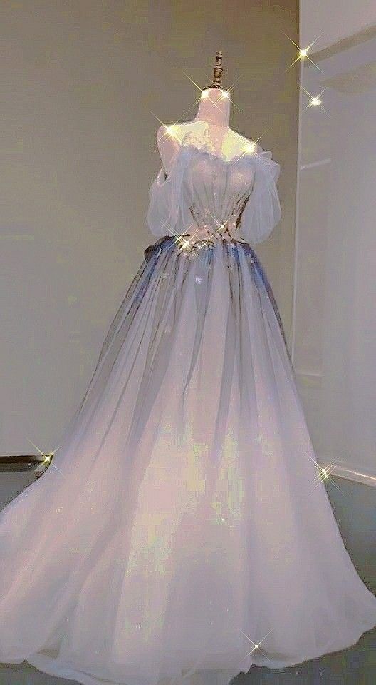 a long, elegant gown is displayed on a mannequin. the dress features an offshoulder design with a delicate bodice, transitioning into a full, flowing tulle skirt in an ombre effect, fading from blue at the waist to white at the hem. it has a fairytale, princesslike aesthetic, with a subtle sparkle adding to its ethereal charm.