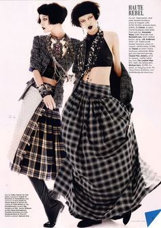 two fashion models are standing in a studio setting. they are both wearing plaid skirts and black tops. the model on the left is wearing a black jacket and has her hair pulled back in a ponytail. the model on the right has her hair down in a straight style. they are both looking towards the camera, and their poses are sophisticated and stylish.  the magazine page that they appear on is styled to give a high fashion vibe, with a headline that reads "haute rebel".  the models are wearing accessories including necklaces, bracelets, and rings.  the fashion style is modern and edgy with a punk inspired aesthetic.