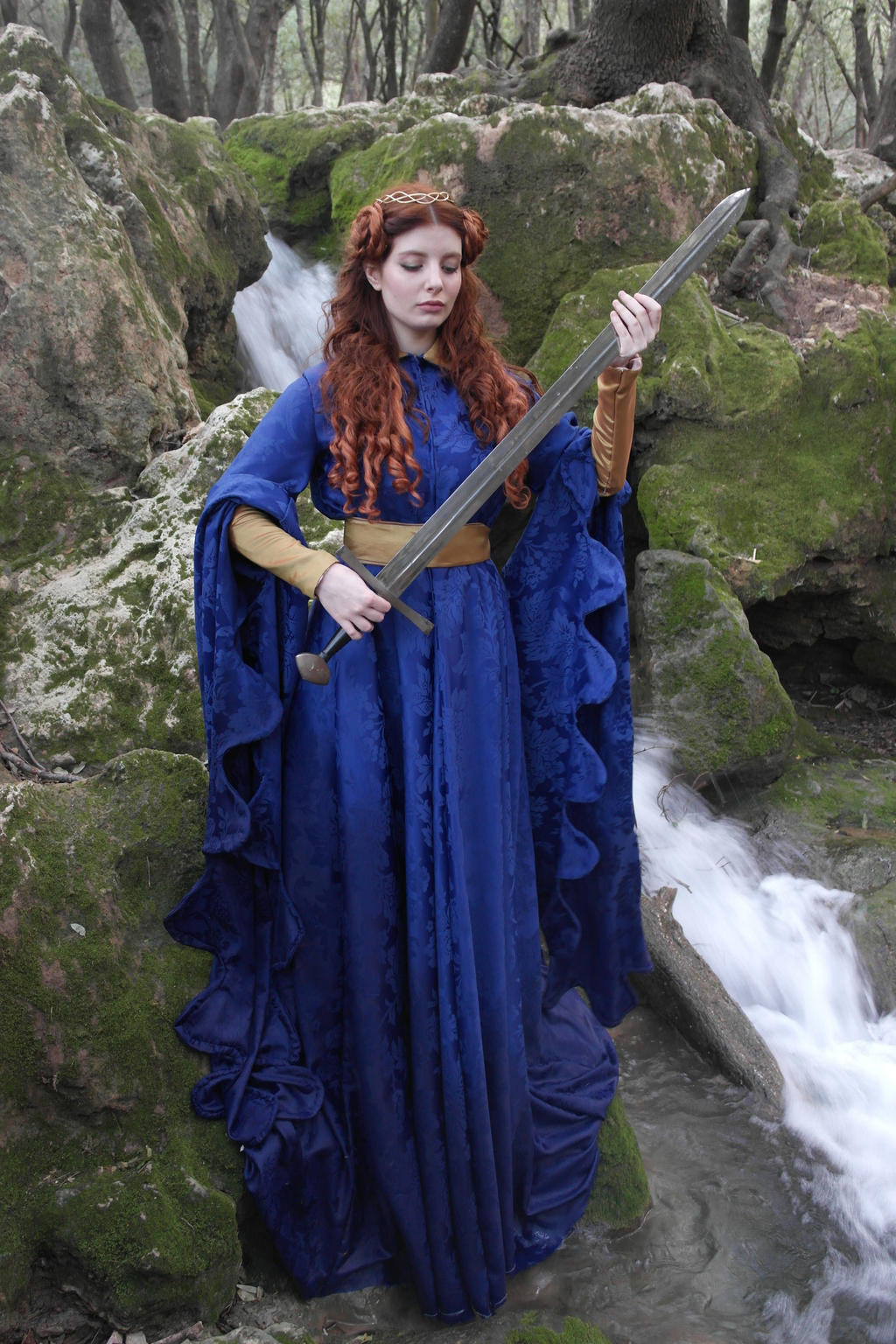 a woman in a long blue medievalstyle dress and a gold crown stands beside a waterfall in a forest. she is holding a large sword, and her expression is calm and serene. the scene is evocative of fantasy or fairytale imagery.  the rocks and mosscovered environment provide a natural, mystical backdrop.  the woman's pose and attire suggest strength and regalness.