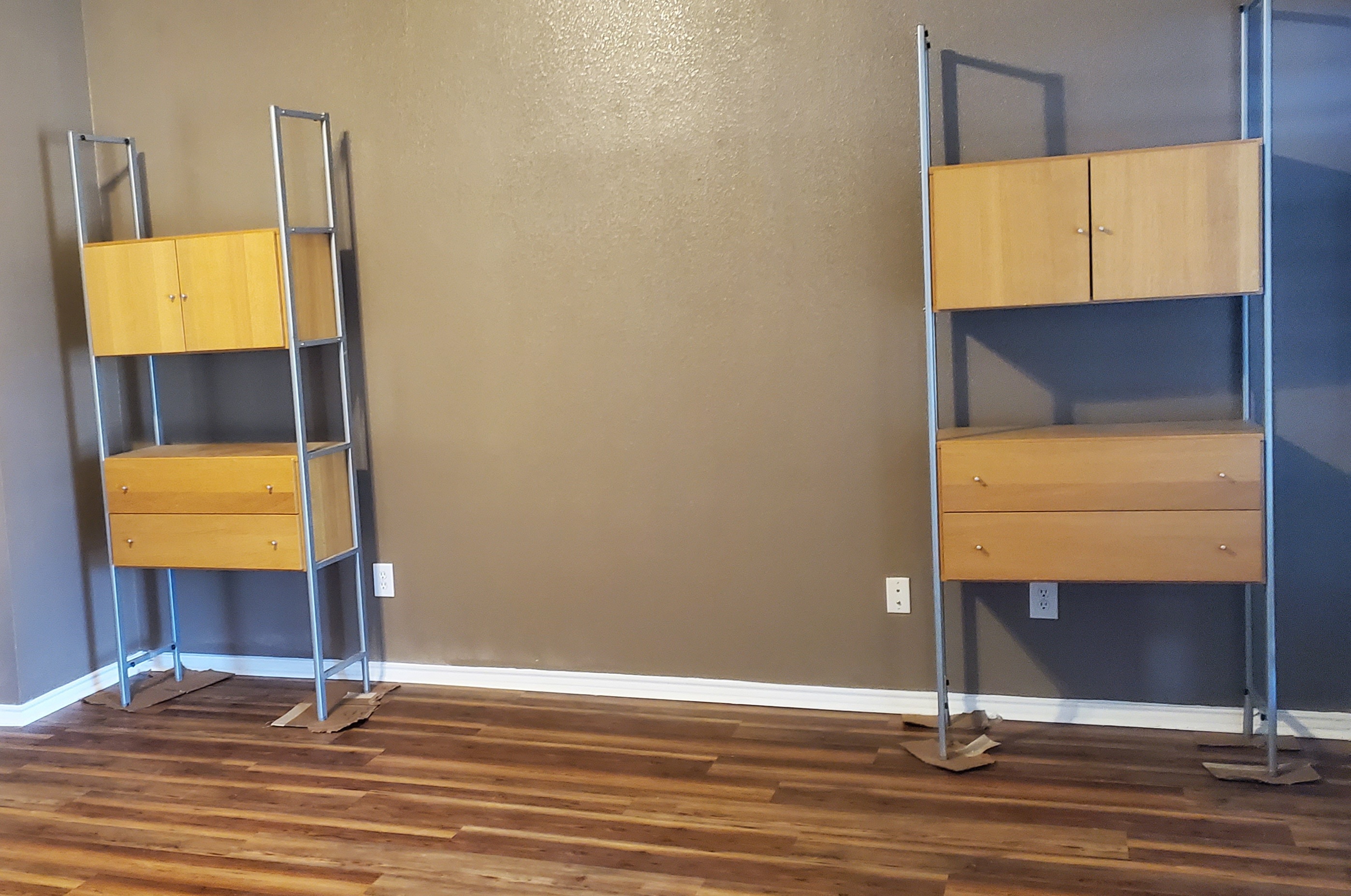 two shelving units with wooden drawers and cabinet sit in a room with brown walls and a wooden floor. the units have silver metal frames. there is an electrical outlet on the wall behind each unit.