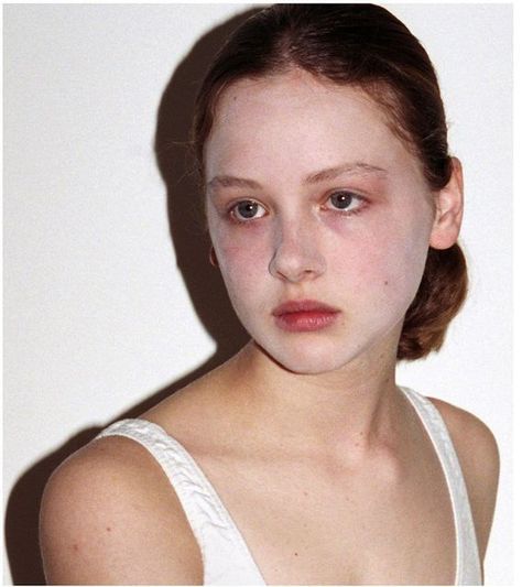 the image is a closeup portrait of a young woman with pale skin and very light makeup.  she has light brown or auburn hair pulled back in a simple bun. she's wearing a simple, white tank top with thin shoulder straps. the background is plain white, drawing all attention to the subject. the overall impression is one of understated, natural beauty.  the lighting is soft and even, suggesting a professional studio photograph.