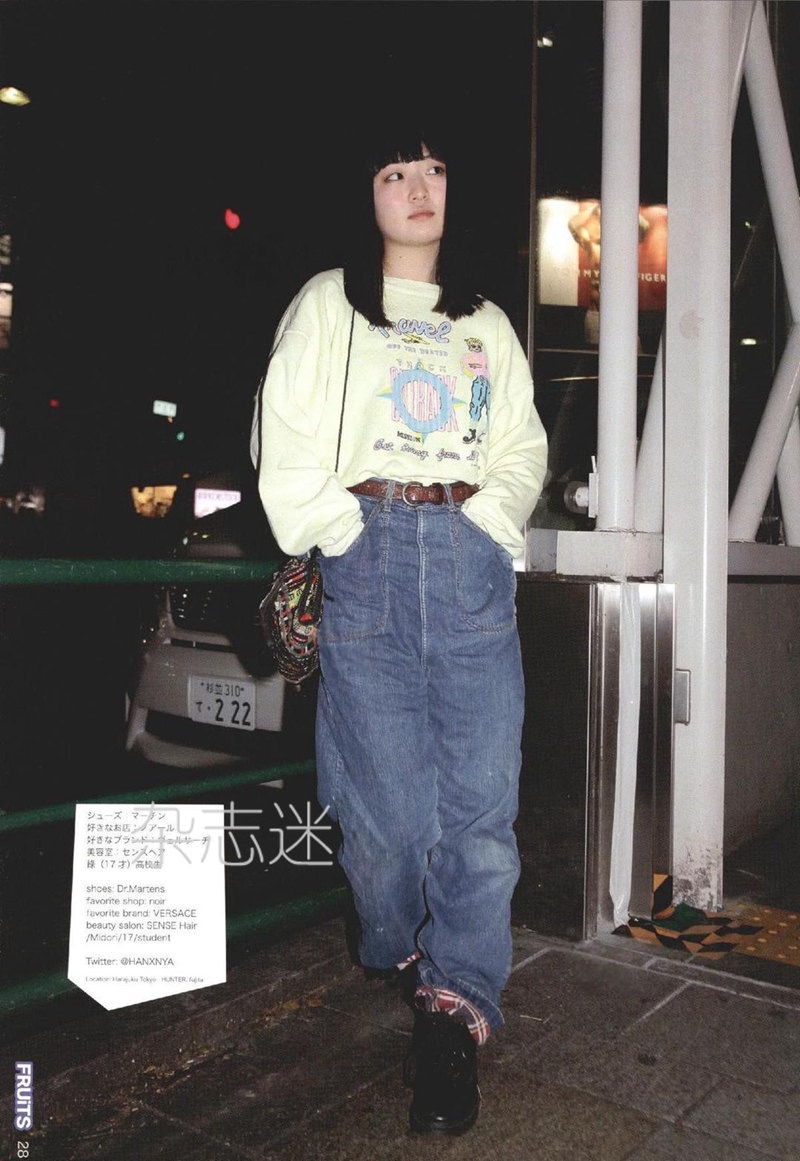 a young woman is captured in a vintagestyle photo, showcasing a 90s japanese street fashion look.  she's posing on a city sidewalk at night, lit by artificial light. she wears an oversized, light yellow sweatshirt with a graphic design, paired with baggy, highwaisted jeans.  her look is completed with platform shoes, a small shoulder bag, and a brown belt. the photo appears to be a scan from a magazine, as indicated by the text and logo at the bottom of the image. ```