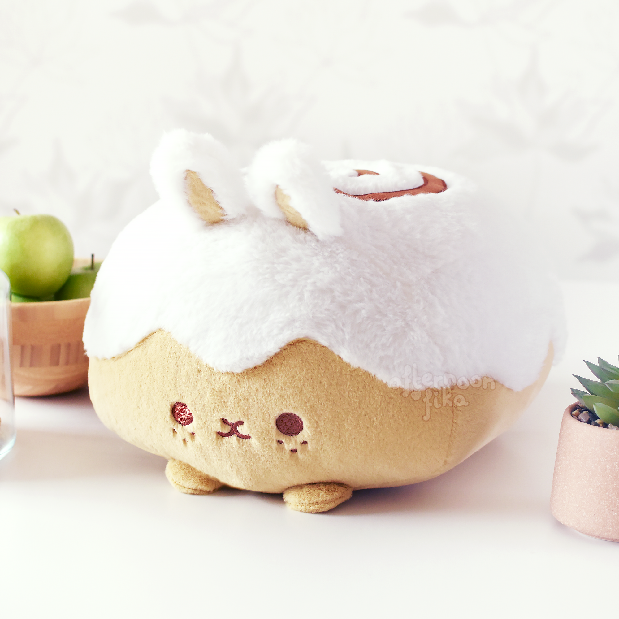 a closeup shot of a handmade cinnamon roll bunny plush toy. the plush is light brown with white frosting and bunny ears. it is sitting on a white surface next to a wooden bowl with apples and a succulent in a pot. the background is a blurry white wall.