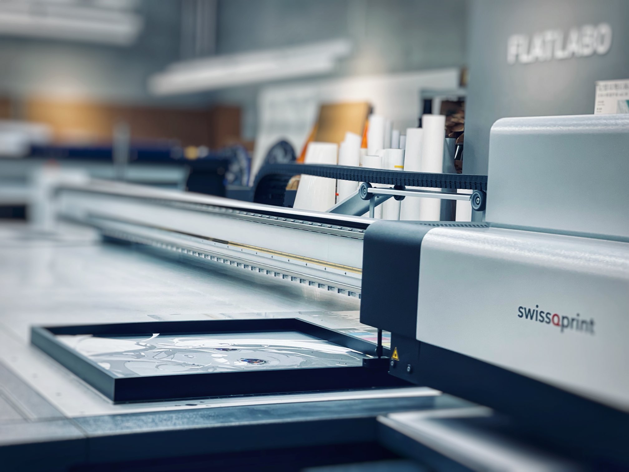 a closeup view of a large format flatbed printer in operation. the printer, a swissqprint model, is creating a print on a rigid material, likely a sign or display graphic. the image highlights the precision and technology involved in modern printing processes. the environment is bright and clean, characteristic of a professional print shop.