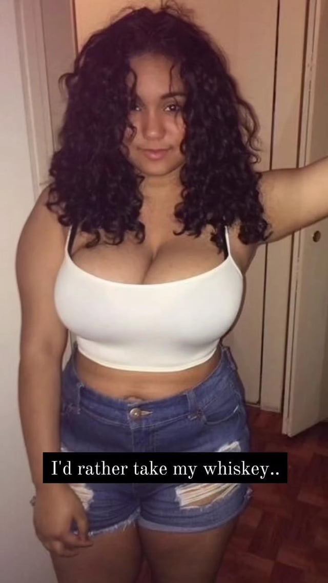 a closeup image of a young, attractive woman with long, curly brown hair. she is smiling and looking at the camera. she is wearing a white crop top and distressed denim shorts. the background is blurred, but it appears to be indoors in a home. a text overlay says "i'd rather take my whiskey...".