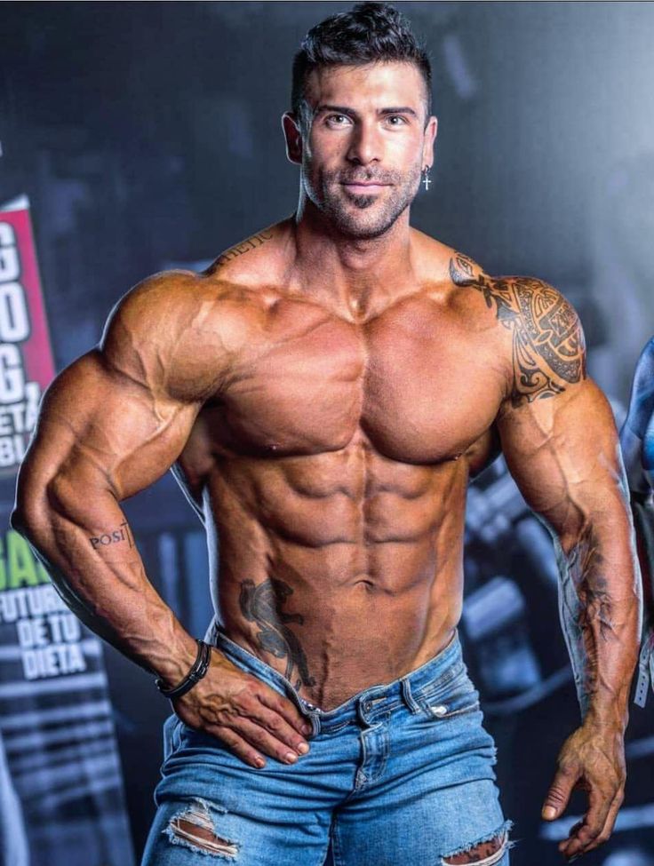 the image showcases a muscular male bodybuilder posing for a studio shot. he has a highly defined physique, with visible abs, veins, and muscular definition throughout his upper body and torso.  he is tanned and has short dark hair,  several tattoos including a prominent tribal design on his right shoulder and a pantherlike design on his left side. he is wearing ripped blue jeans and a small cross earring. the background is blurred, but suggests an indoor setting possibly related to a fitness competition or photoshoot. the overall impression is one of strength, fitness, and athleticism.