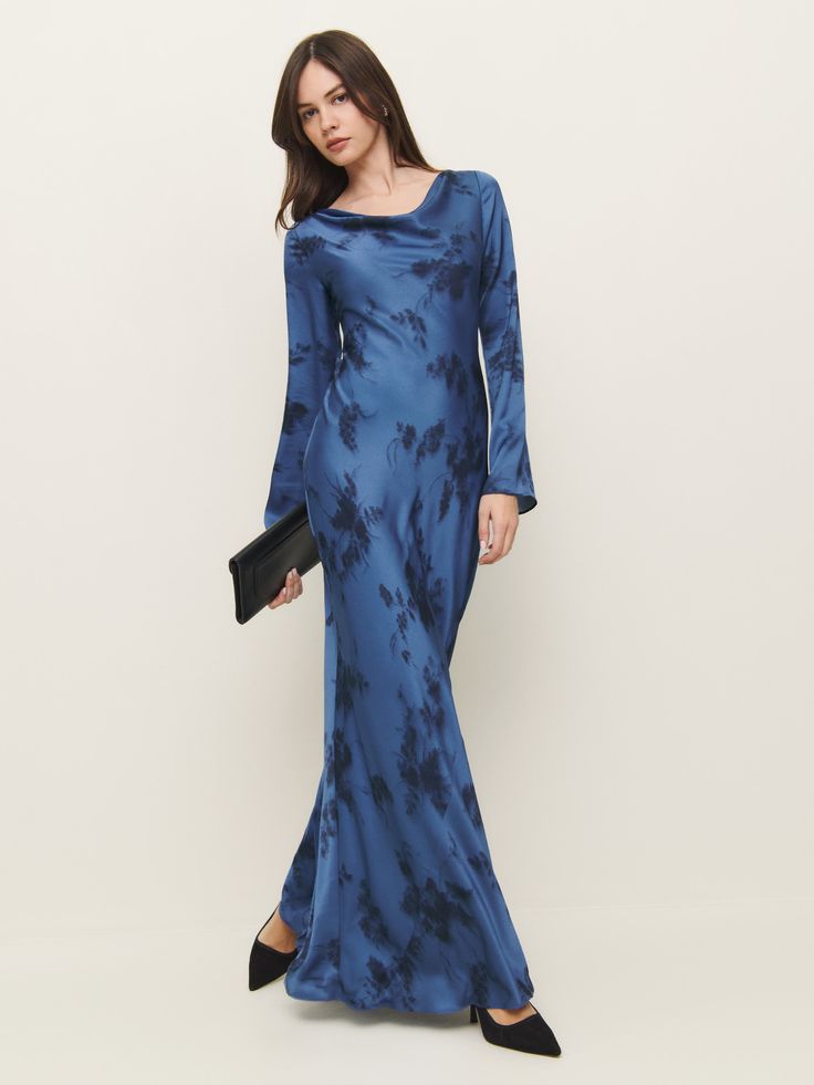 a fulllength studio shot of a woman wearing a blue floral maxi dress. the dress is a slipstyle dress made of satin material with long sleeves and a cowl neckline. the woman is wearing black heels and carrying a black clutch. the background is a simple white studio backdrop.