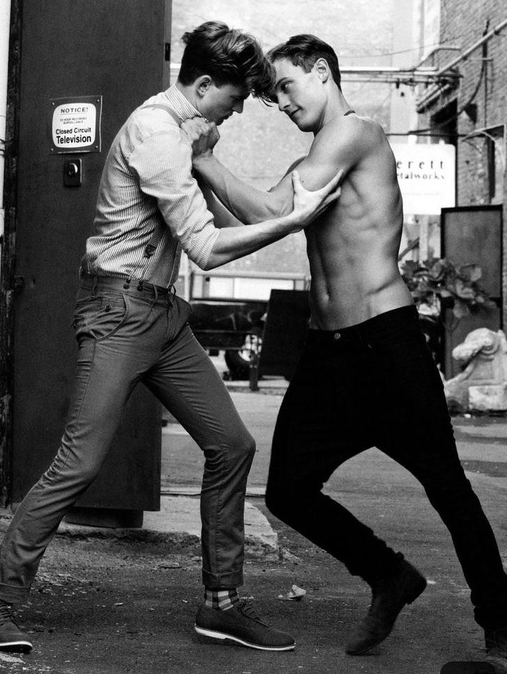 two young men in an urban setting, one shirtless with a muscular build and the other wearing a shirt and pants, are seen fighting on a city street.