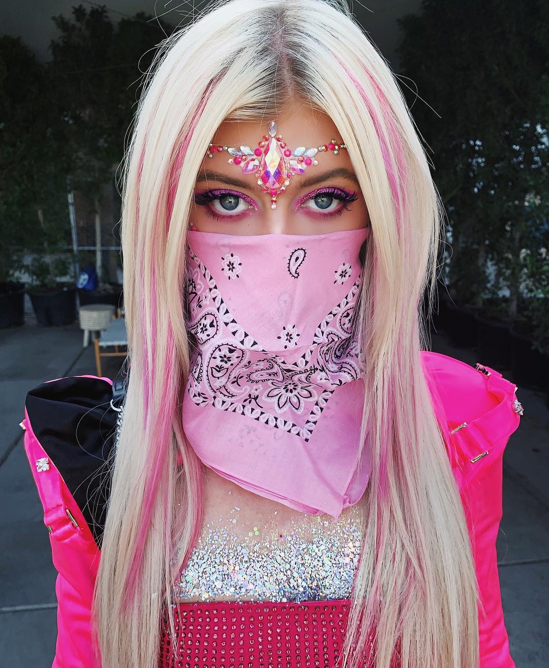 a woman with blonde hair and pink highlights is wearing a pink bandana over her face. she has face jewelry on her forehead and is wearing glitter on her chest. she is also wearing pink eyeshadow and a pink jacket.
