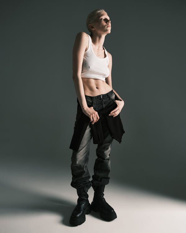 a fulllength studio shot of a woman with short, light blonde hair posing in a fashionforward outfit. she is wearing a cropped white tank top that reveals her midriff. her pants are lowrise, faded black jeans, and she has a black jacket tied around her waist. she is wearing chunky black boots. the background is a plain grey, and the lighting is focused on her, highlighting her figure. she is looking up and to the side with a confident expression.