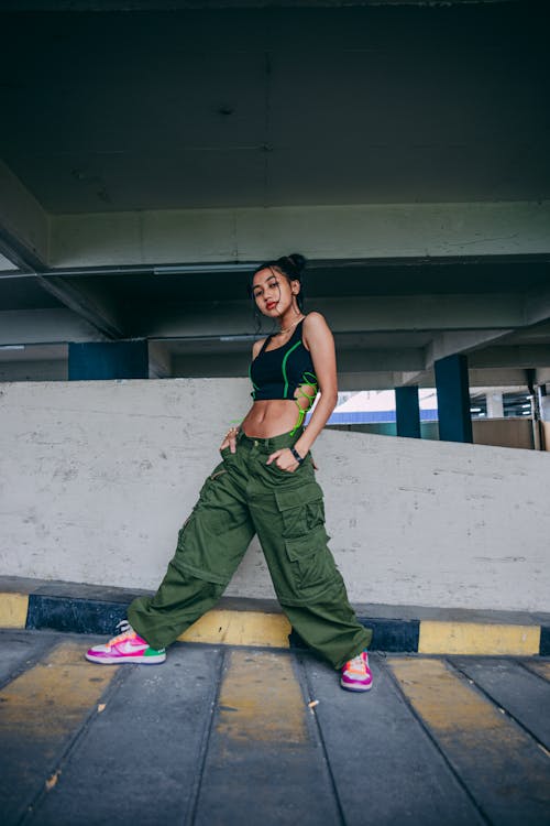 a young asian woman poses confidently in a parking garage. she is wearing a black crop top, olive green cargo pants, and pink and white sneakers.  her style is urban and trendy. the setting is industrial with a concrete wall and floor. she is looking directly at the camera.