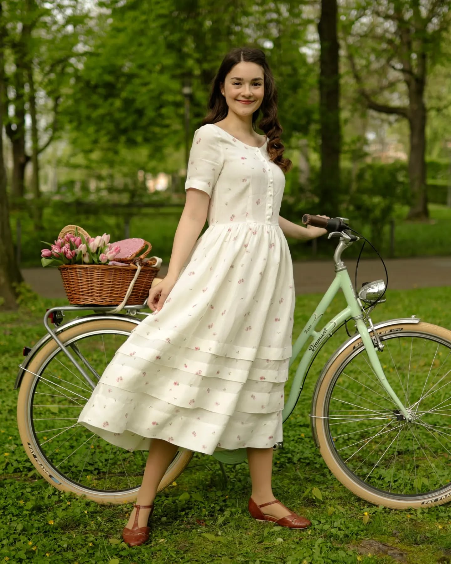 a young woman with long brown hair is smiling happily. she's wearing a vintagestyle, offwhite midi dress with a small cherry print. the dress has short sleeves and a buttondown front. she’s leaning against a pastel green vintage bicycle which has a wicker basket filled with tulips and a picnic basket attached to the front. the setting is a sunny park with lush green grass and trees in the background, creating an idyllic and romantic atmosphere. the overall style evokes a feeling of nostalgia and vintage charm. the woman is wearing brown mary jane style shoes.