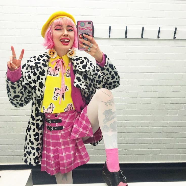 a woman with pink hair and a yellow beret is taking a selfie in front of a brick wall. she is wearing smiley face earrings, a cow print coat, a yellow shirt with a dog print, a pink sweater, a pink plaid skirt, white tights with tattoos, and pink socks with black shoes. she is holding up a peace sign.