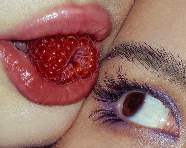 a closeup shot features two individuals in intimate proximity. one person's lips are prominently displayed,  partially obscuring a whole, ripe raspberry. the lips are full, glossy, and a deep pinkishred.  the other person's eye, accented with purple eyeshadow and long dark eyelashes, is visible.  the overall aesthetic is sensual and visually striking, with a focus on texture and color contrast between the bright red raspberry and the purple eye makeup.  the image suggests a theme of beauty, intimacy, and the juxtaposition of food and makeup.