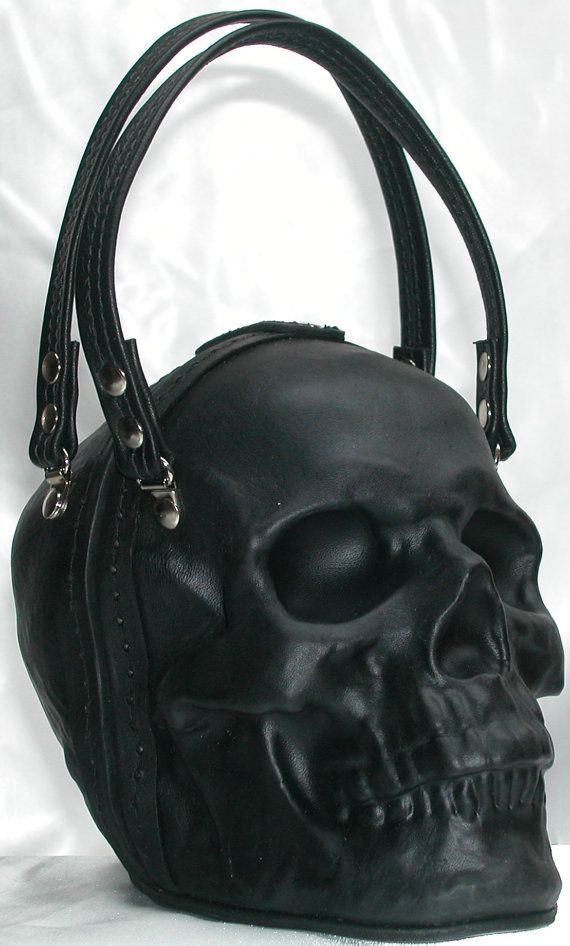 a black leather purse shaped like a human skull is shown. it has two arched handles that connect to the skull via straps with rivets. the skull's features, including the eyes, nose, and teeth, are clearly defined. it is set against a plain background.