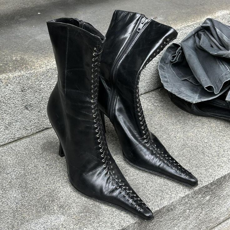 a closeup shot of a pair of black leather stiletto boots with pointed toes and laceup detailing on the sides.  they are positioned on a stone staircase with folded darkcolored clothing resting nearby. the boots are sleek and elegant, showcasing fine details in their design and craftsmanship. the overall image suggests a streetstyle fashion context or a stilllife representation of the footwear.