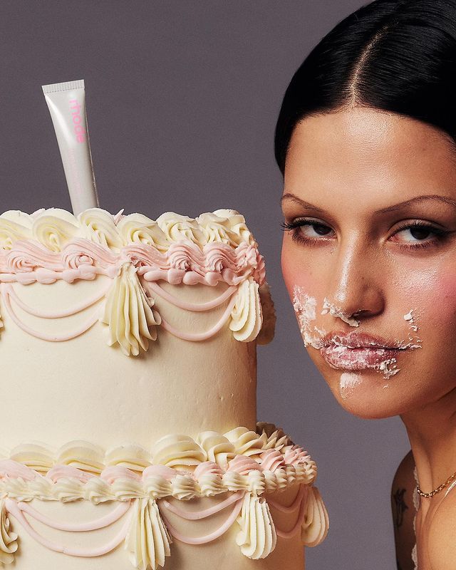a woman with black hair is smiling with a face covered in frosting while looking at the camera.  the woman is leaning into a large, twotier cake decorated with pink and white frosting. she is holding a tube of frosting in her hand.