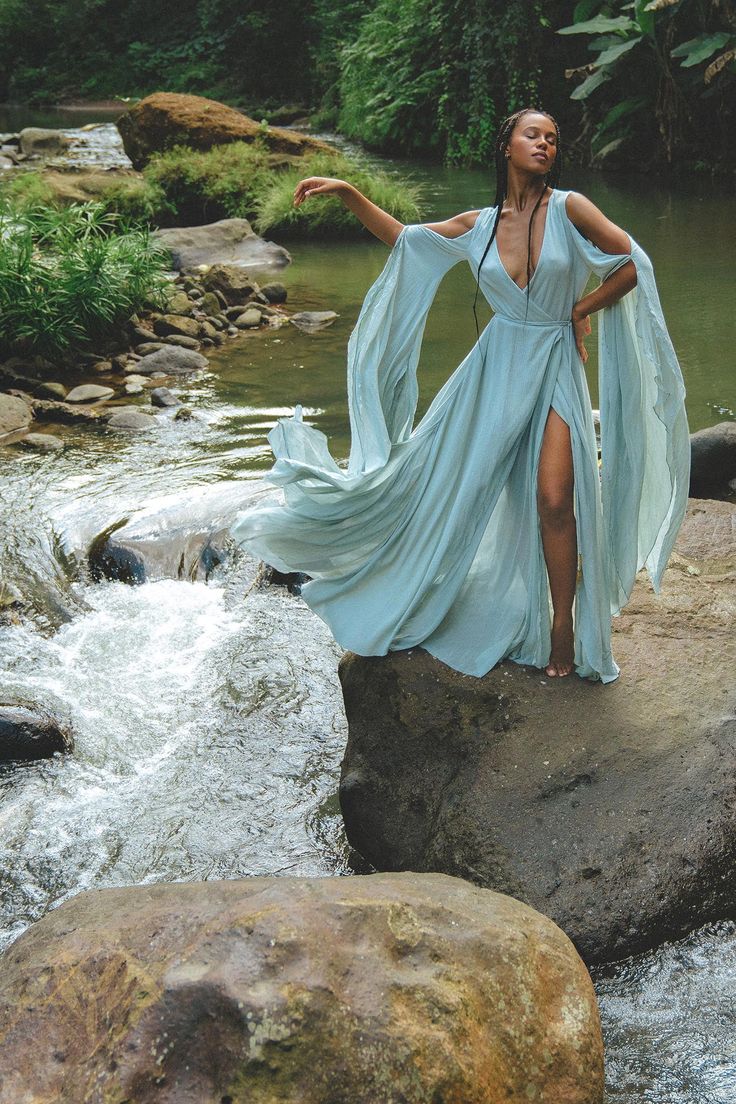 a black woman with long braids stands gracefully on a rock in a flowing, light blue maxi dress with wide sleeves and a thighhigh slit.  the dress billows dramatically in the gentle breeze.  she is positioned amidst a beautiful, lush natural setting beside a flowing river or stream, with rocks and green foliage surrounding her. the overall mood is serene, peaceful, and ethereal. the image suggests a fashion photoshoot or a stylized portrait emphasizing the model's elegance and the beauty of her surroundings.