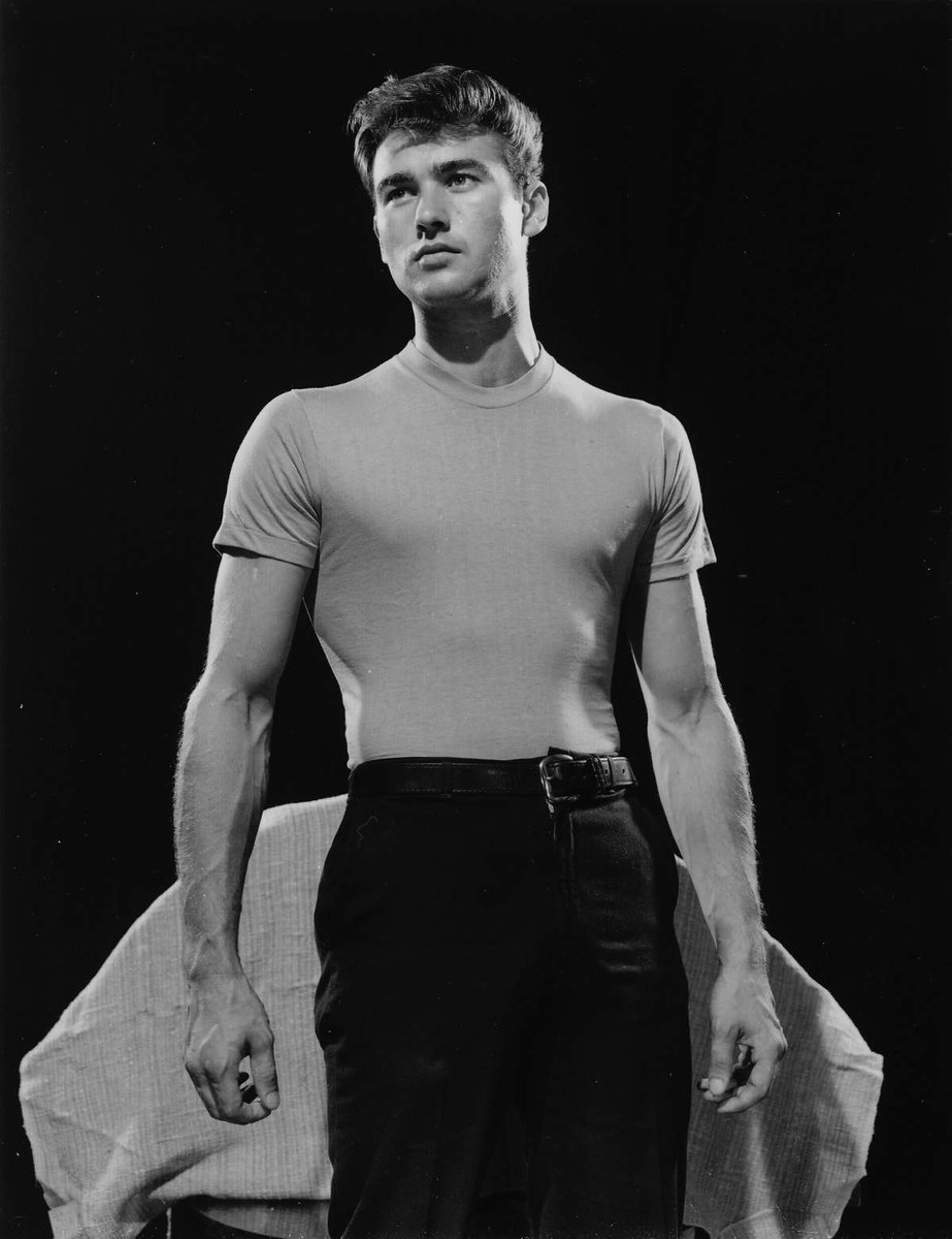 a black and white studio portrait of a young man in a classic 1950s outfit, wearing a fitted tshirt and dark pants with a belt. he stands with his arms slightly away from his body, looking offcamera with a contemplative expression. the dramatic lighting emphasizes his physique and features, creating a sense of timeless cool.
