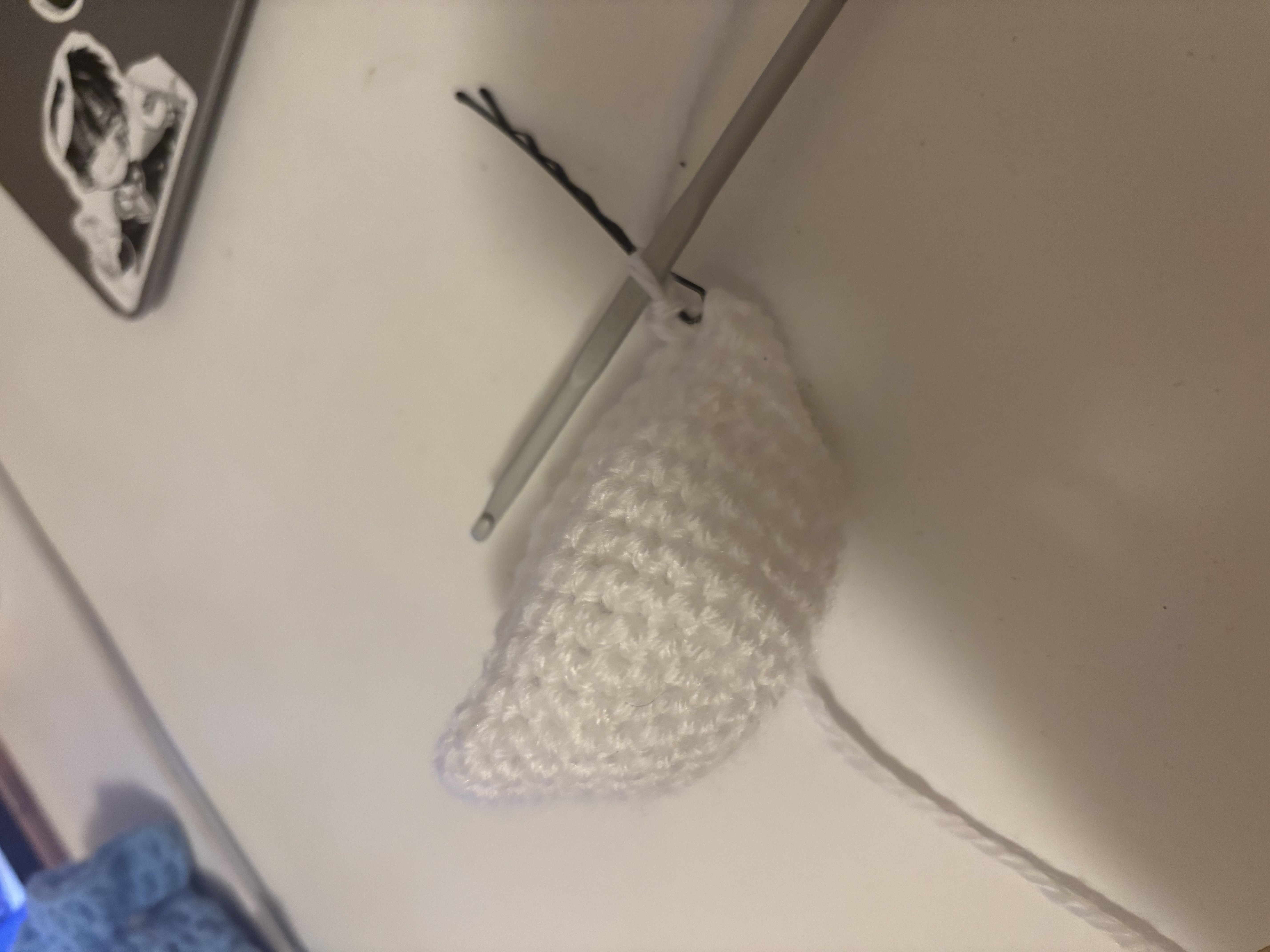 a closeup shot shows a partially crocheted white amigurumi piece with a crochet hook inserted. a white strand of yarn extends from the crocheted piece. to the right of the crochet piece is a black hairpin. in the background, part of a laptop with a sticker on it is visible. the background is a lightcolored surface.