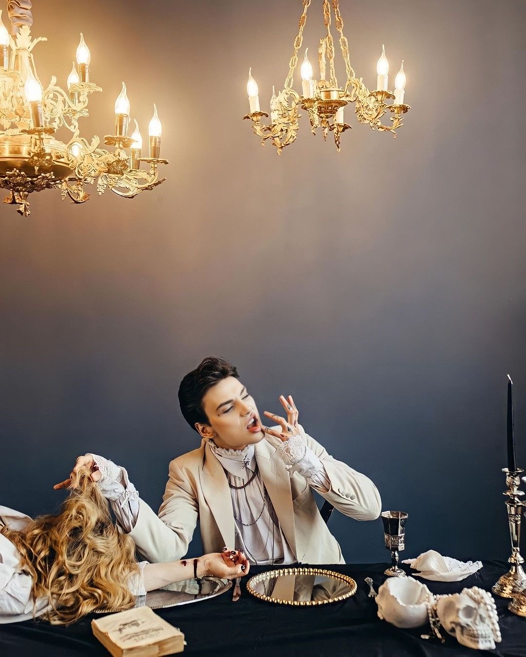 in a dimly lit room, a man and a woman are positioned at a table set for a spooky feast. the scene evokes a vampire theme with gothic and victorian elements. the man, dressed in a white victorianstyle shirt,  reaches towards his mouth with two fingers as if tasting something. his eyes are closed, and his expression suggests he's savoring a rich, perhaps bloody, flavor.  the woman is draped across the table, her long blonde hair cascading down, and one hand reaching towards the man.  her face is not visible. the table is adorned with candles, a skull, and antiquelooking goblets, amplifying the eerie and dramatic atmosphere. two ornate chandeliers illuminate the scene, casting a soft, warm glow against the dark background.