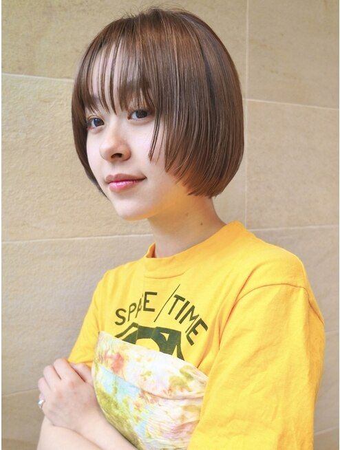 a woman with short brown hair and bangs is looking at the camera and smiling slightly. she is wearing a yellow tshirt with a graphic on the front. the background is a white wall with a light tan tiled wall. the image is a closeup portrait.
