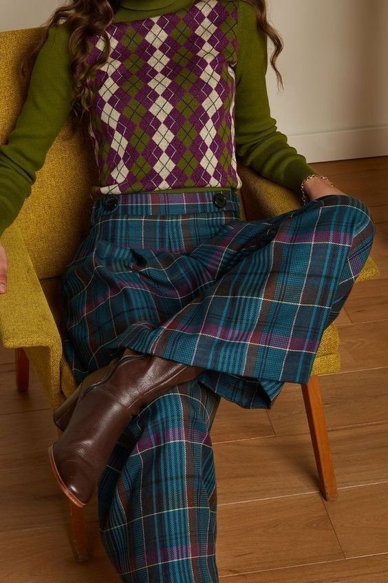 a woman is sitting on a chair with a green argyle sweater, blue plaid pants, and brown boots. the chair is yellow, and the floor is wooden. the woman is wearing a classic 70s or 80s style outfit. she is looking down, and her pose is relaxed.
