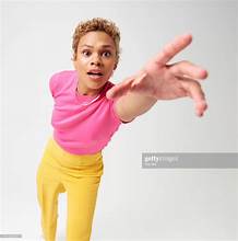 a woman with short hair styled up is wearing a pink tshirt and yellow pants, reaching out towards the camera with a surprised expression on her face. she is set against a plain white background.