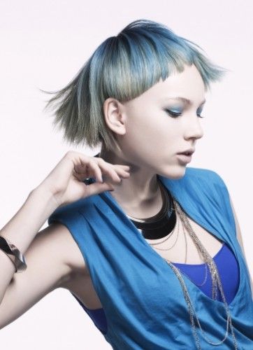 a young woman with blue and green short hair is wearing a blue top with silver necklaces and a black choker. she is looking to the side with a slight smirk. the image is well lit and the model is posed against a white background.
