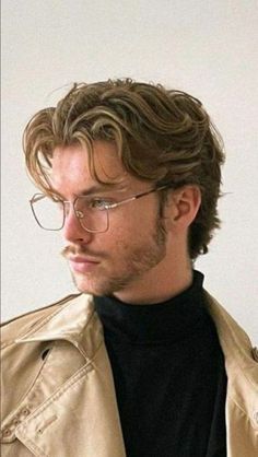 a man wearing glasses, a black turtleneck, and a tan trench coat is pictured in a closeup. the man has blond, wavy hair and a beard and mustache. he is looking to the side.