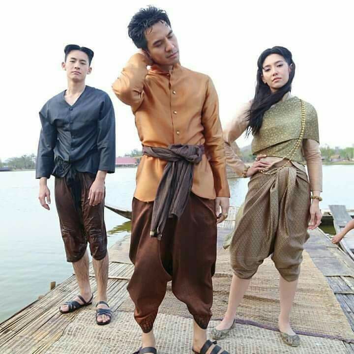 a group of three people in traditional thai clothing stand on a dock. the men are wearing longsleeved shirts and pants while the woman is wearing a wrap skirt and a blouse. they are all smiling and looking happy.