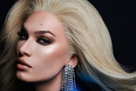 the image is a closeup portrait of a drag queen, likely a professional model, with striking features and makeup. she is wearing a long, flowing blonde wig styled in a dramatic fashion that sweeps across her shoulders and down. her makeup is heavy but expertly applied, featuring a smoky eye look and highlighted cheekbones and skin.  she also sports large, dangling crystal earrings.  the overall mood is one of glamour and high fashion. the background is a simple, dark backdrop which keeps the focus firmly on the subject.