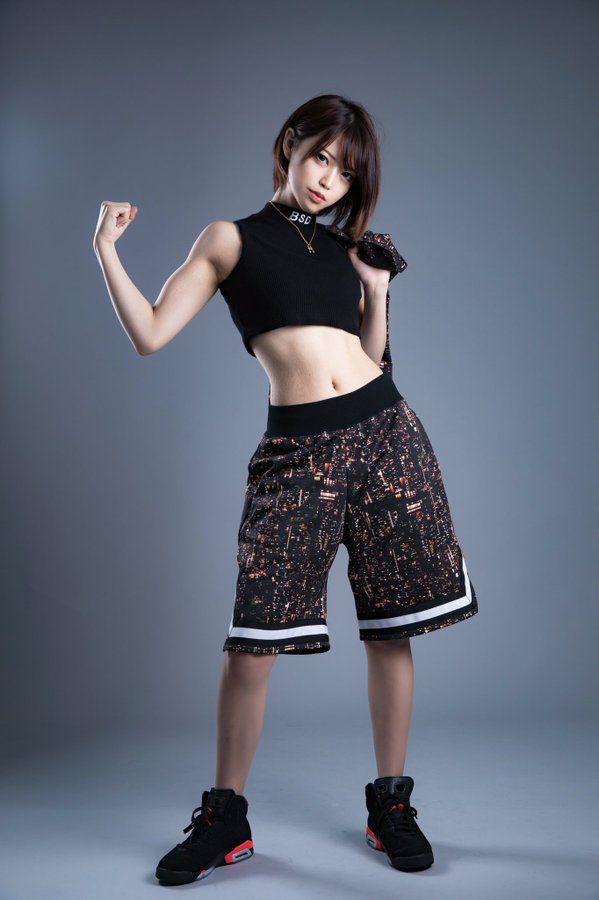 a young asian woman, possibly a model, is posing in a studio against a gray backdrop. she's dressed in athletic attire: a black highnecked crop top, patterned basketball shorts, and black sneakers with orange accents. her short hair is styled simply.  she's flexing one bicep, showcasing a confident and healthy physique. the overall style is sporty and trendy.