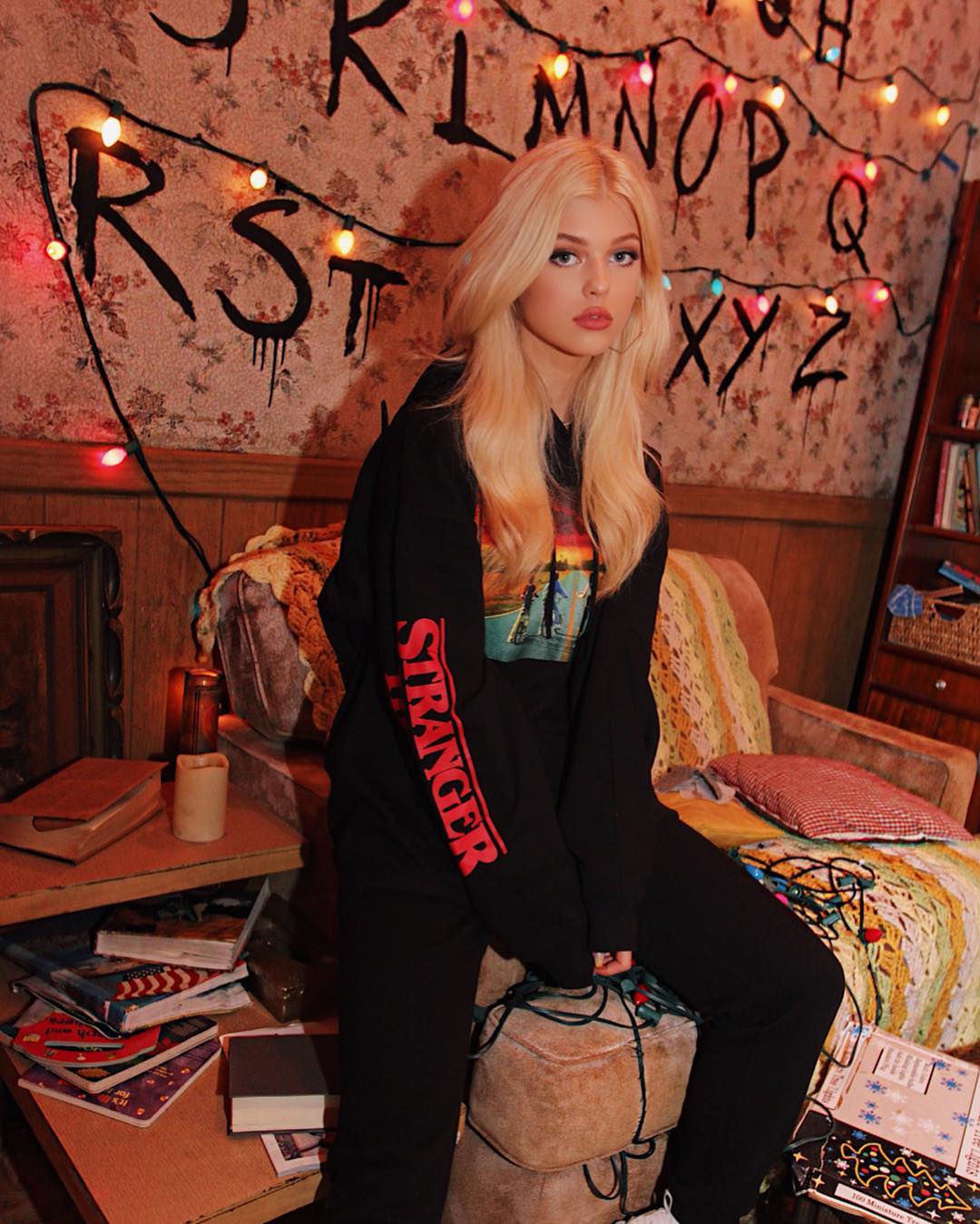 a young woman with long blonde hair, loren gray, is sitting in a room decorated in a "stranger things" theme. she is wearing a black sweat suit with red "stranger" letters on the sleeve and the room has alphabet letters written on the wall, draped with christmas lights. the room also features a vintage chair, books on a table and other cozy decorative elements.