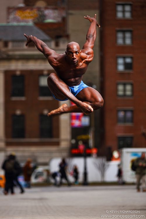a man in a blue and white striped dance leotard jumps in mid air with his arms and legs extended in a contemporary dance pose in front of a building.  the background is slightly blurry but shows other people walking on the city street.