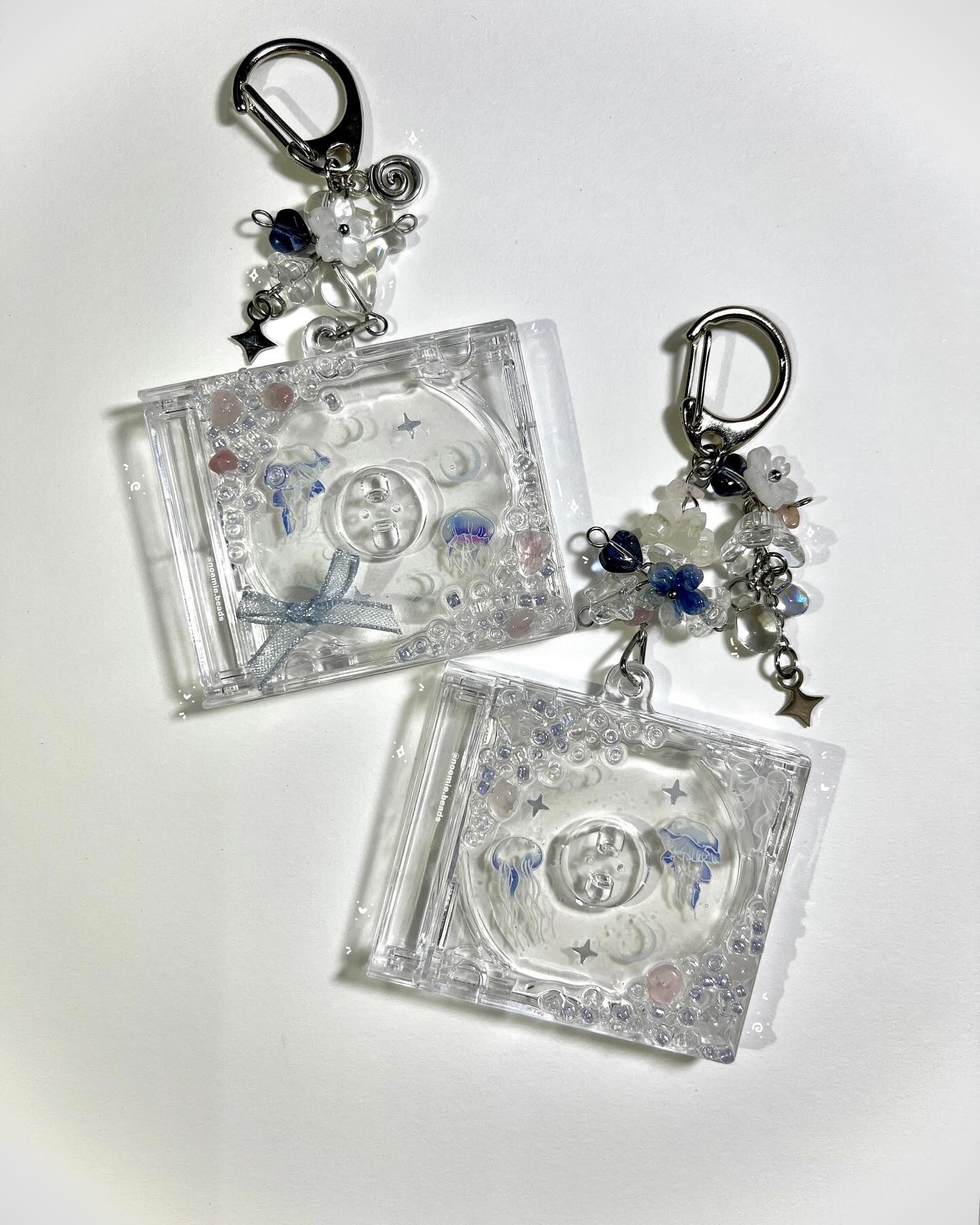 the image shows two handmade keychains. each keychain consists of a clear, square cd case decorated with small beads in pink and blue, tiny white flowers, and miniature silver stars. inside the cd case, there is a small replica of a cd that also features blue jellyfish designs and stars. the cd cases are attached to silver clasps with beaded and floral embellishments, giving the keychains a whimsical and handmade look. the keychains are on a white background.
