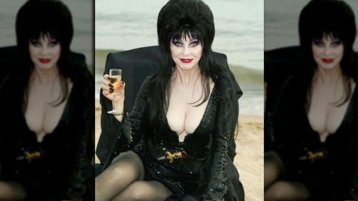 a woman in a black dress with a plunging neckline sits on a chair, holding a glass of champagne and looking into the camera. her dark hair is styled in a dramatic updo, and she appears to be enjoying herself.
